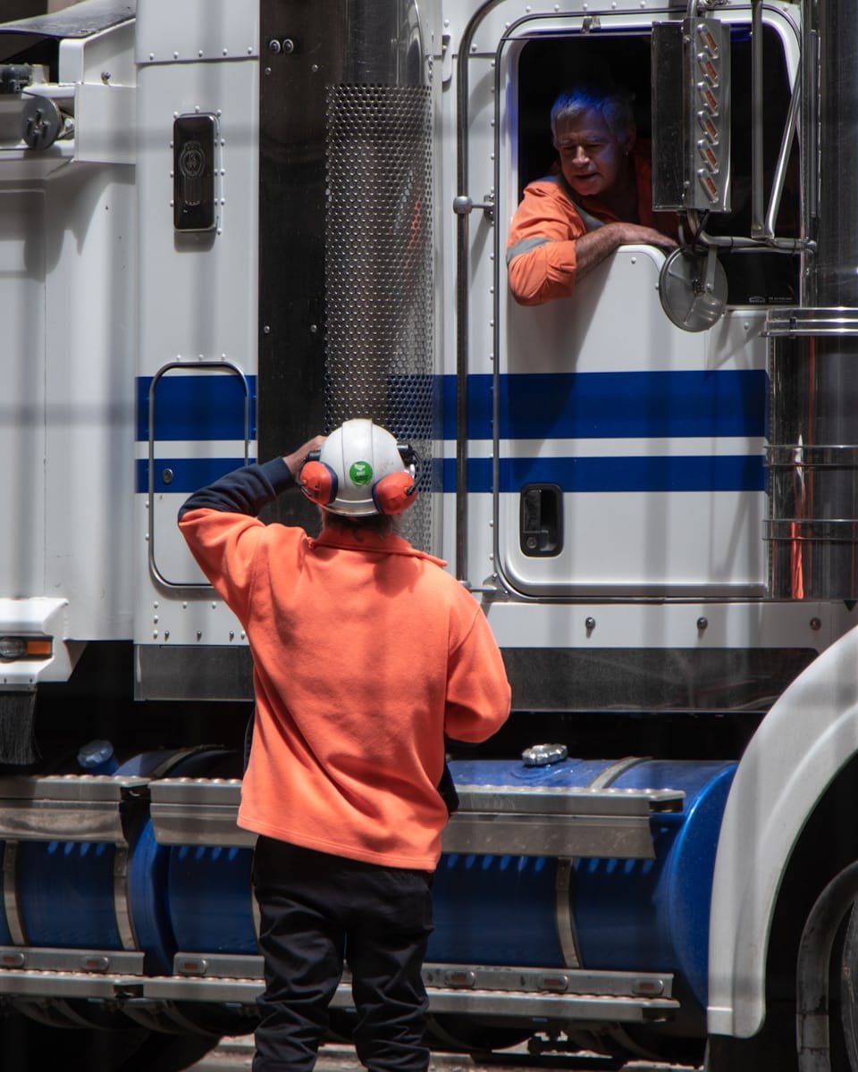 An instructor in high-visibility clothing talking to a driver seated in a truck cab.