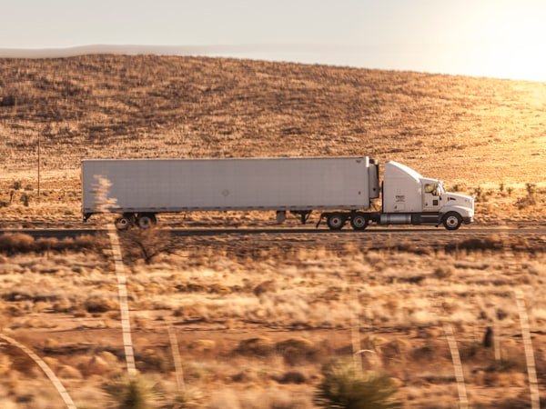 A prime mover and trailer travelling at speed along a highway through dry scrub.