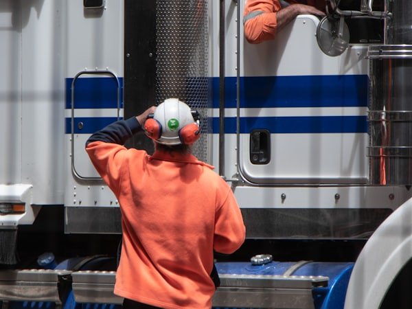 A worker in high-visibility clothing speaking to a driver seated in a truck cab.