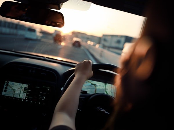 View over a driver's shoulder with hands on the steering wheel, looking down a road at dusk.